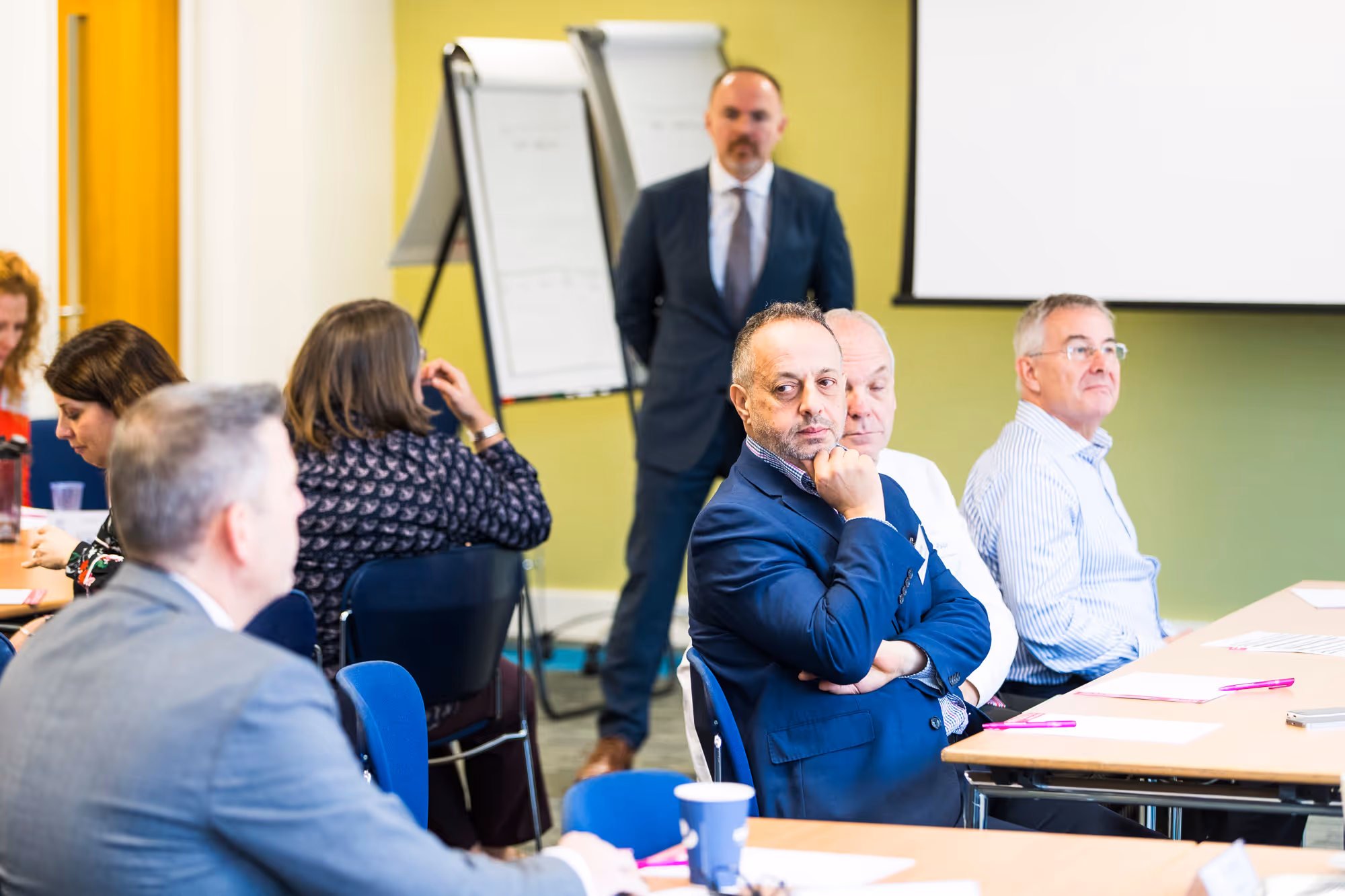 People seated around tables in a meeting or workshop room with a man standing near flipcharts in the background.
