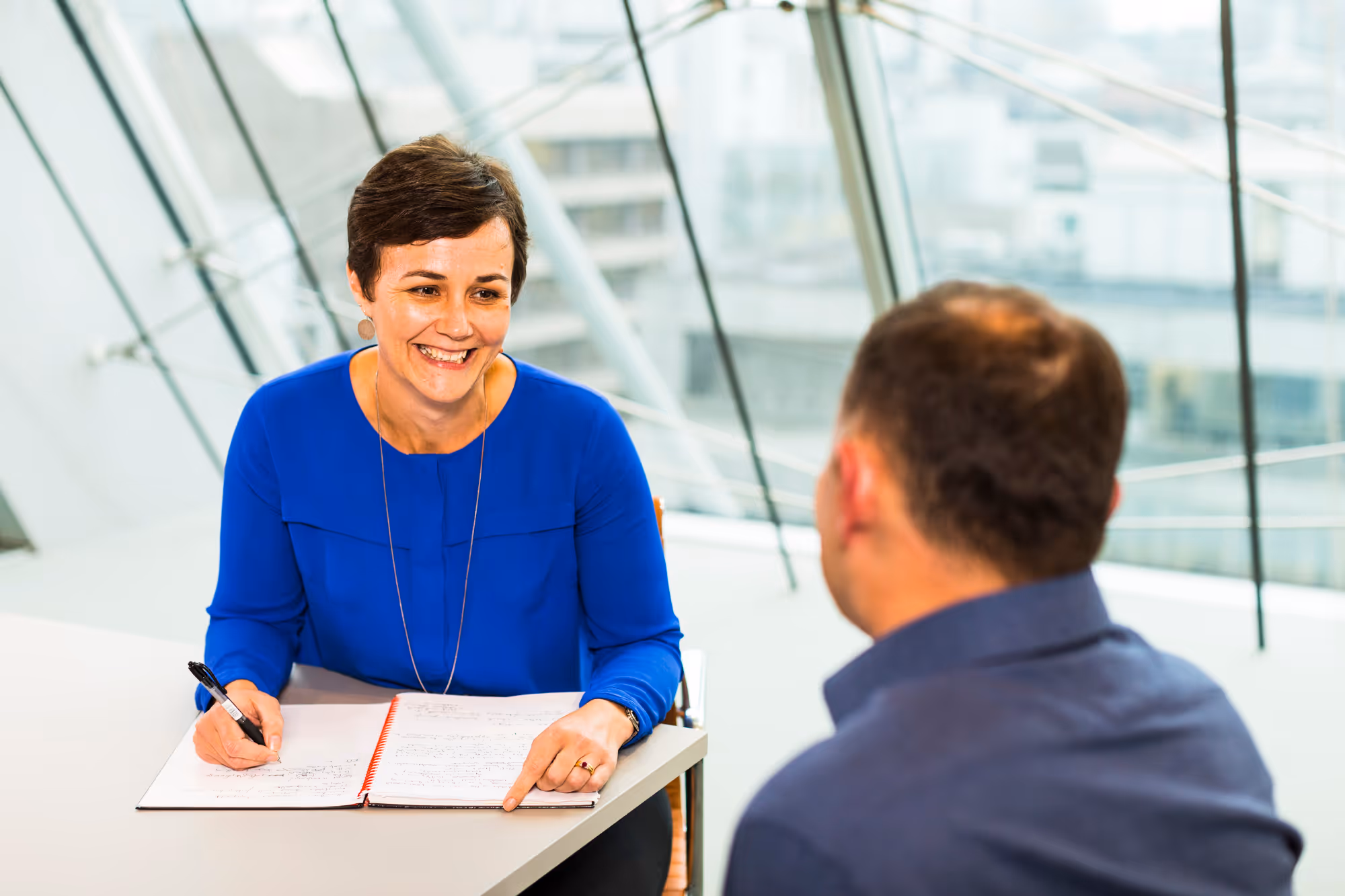Smiling woman in blue shirt holding a pen and pointing at a notebook during a conversation with a man.