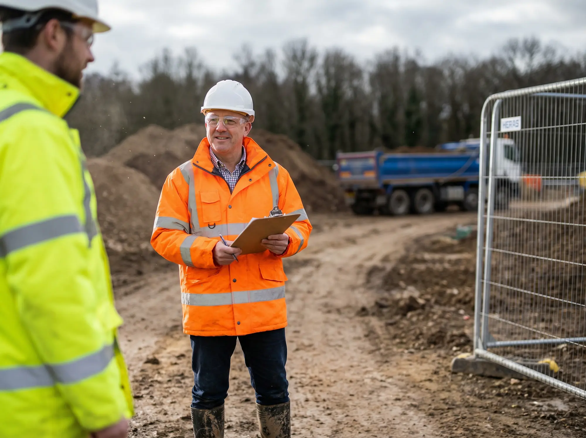 Construction worker in orange safety jacket and hard hat holding a clipboard and talking to another worker in yellow jacket at a muddy construction site with a truck and fencing in the background.
