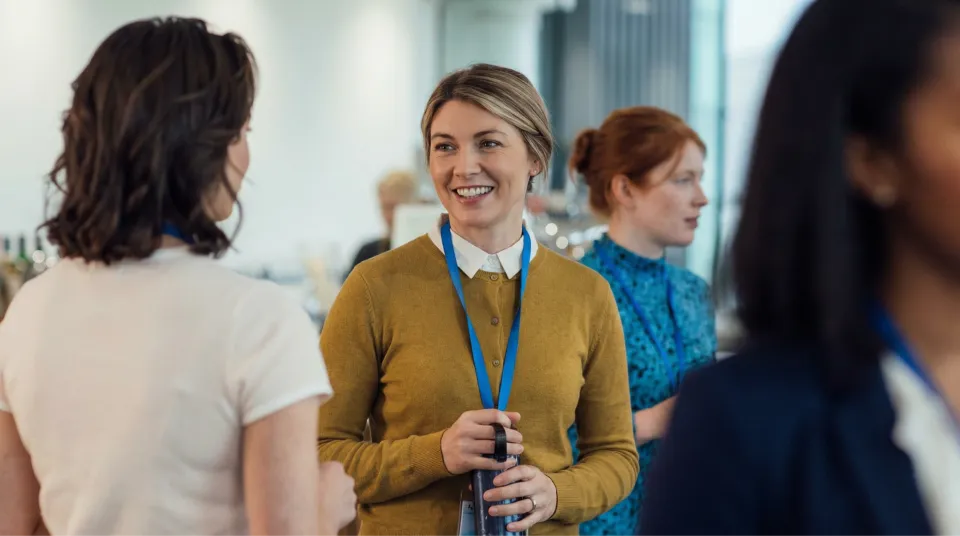 A smiling woman in a mustard cardigan and white collared shirt holds a water bottle and wears a conference badge, talking to another woman.