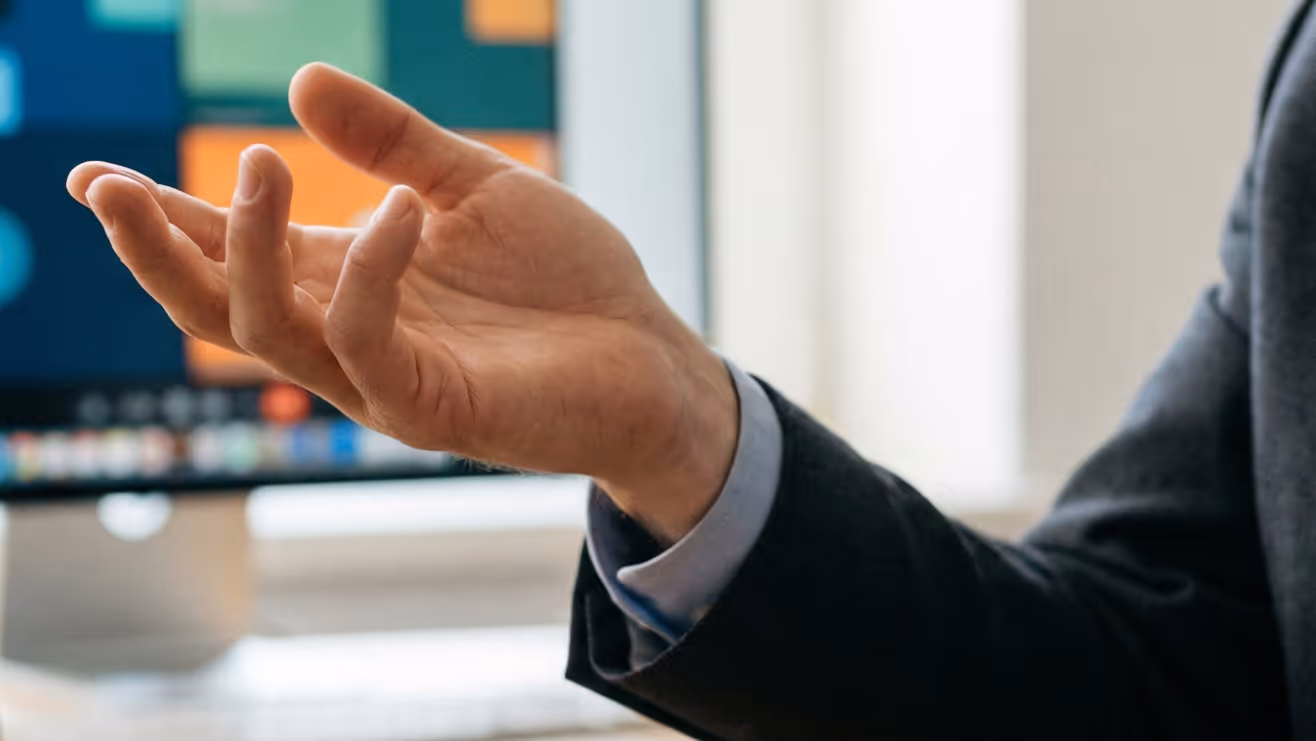 Close-up of a person's hand gesturing with an open palm in front of a blurred computer screen.