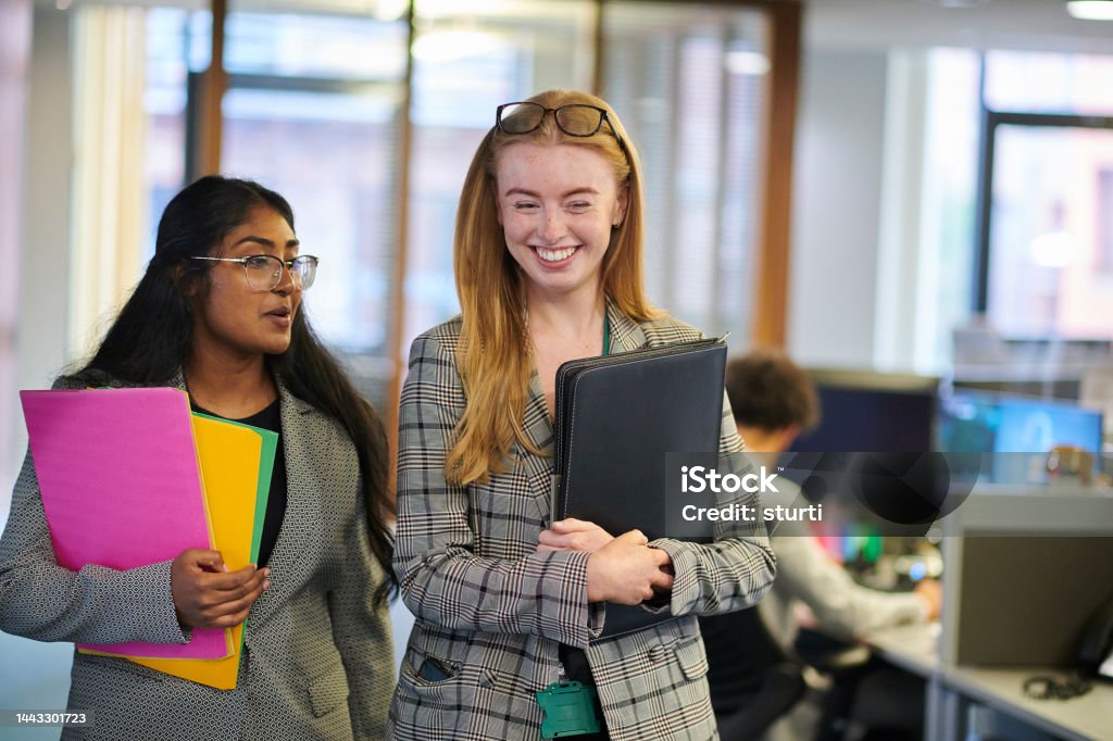 Two young professional women walking indoors while holding colorful folders and a black portfolio, smiling and talking.