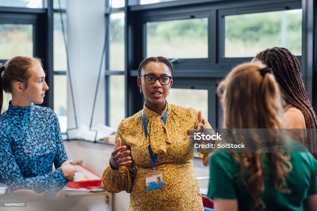 A woman in a mustard yellow blouse speaking and gesturing to three attentive women in a bright room with large windows.
