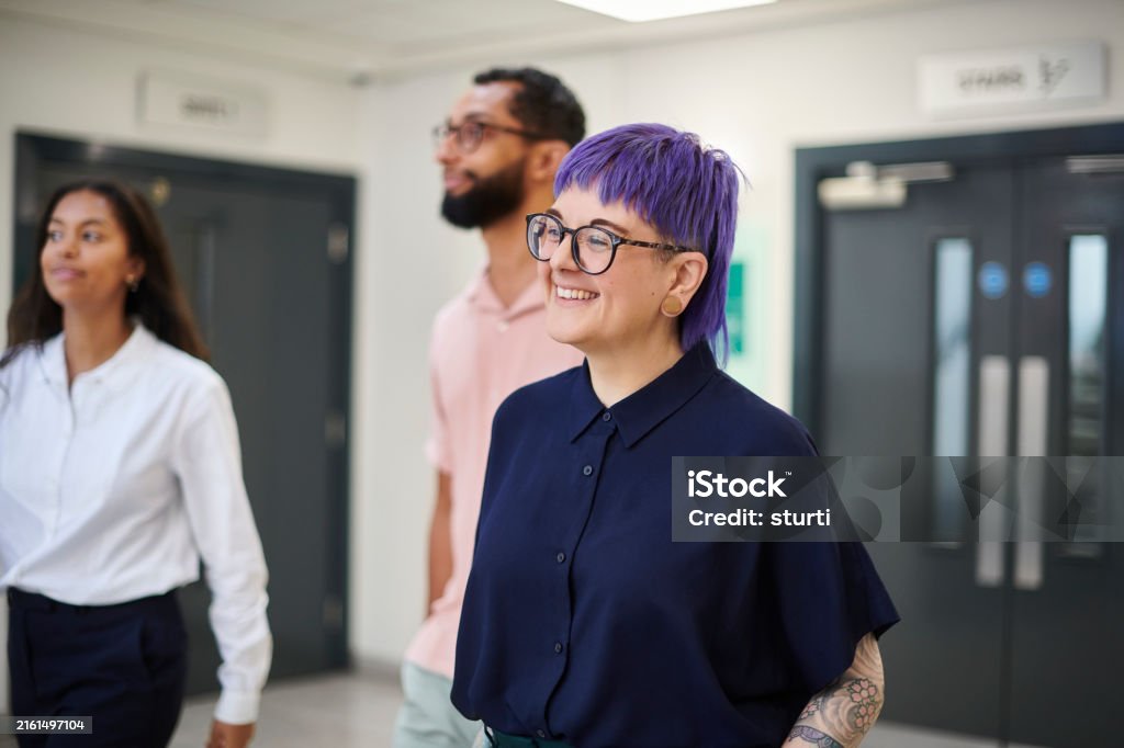 Three young adults walking indoors, one with purple hair and glasses smiling in the foreground.