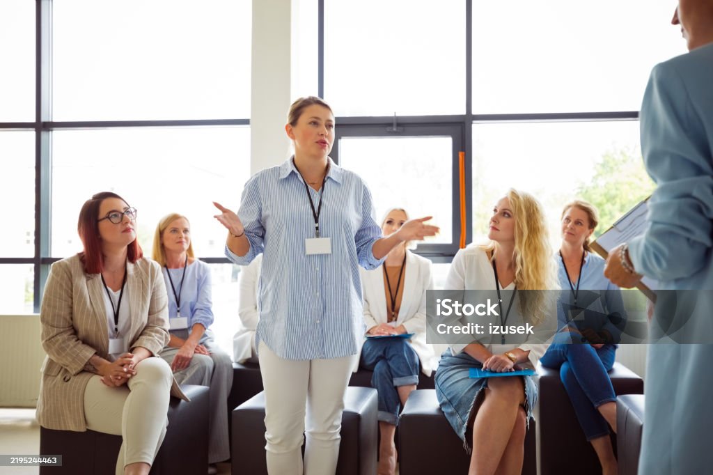 Woman in a blue striped shirt standing and speaking to a group of seated women attentively listening in a bright meeting room.