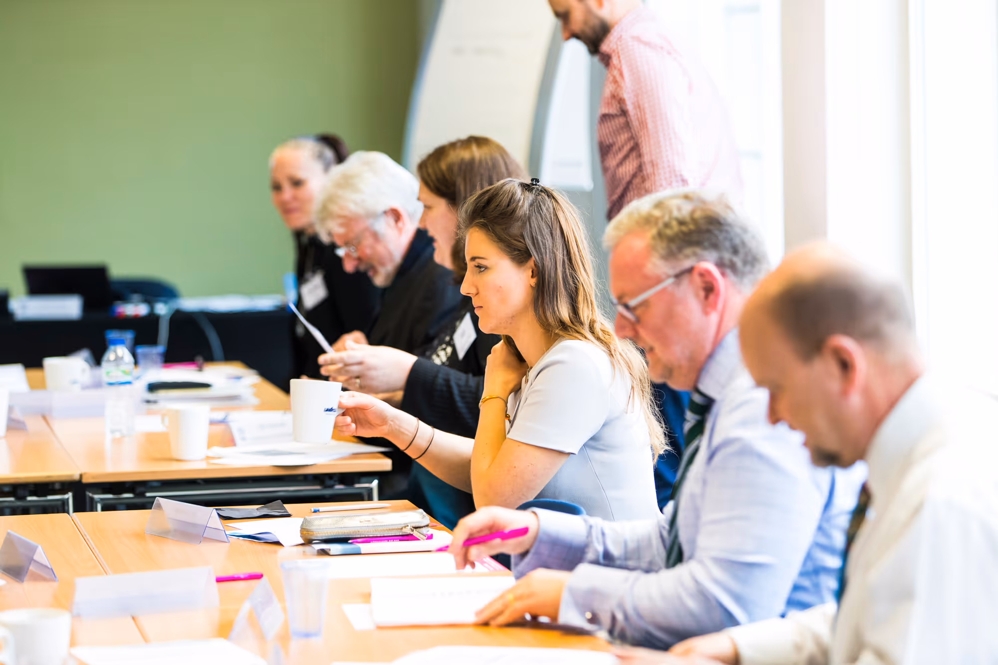 Group of people sitting at a conference table reviewing documents and holding coffee mugs in a meeting room.