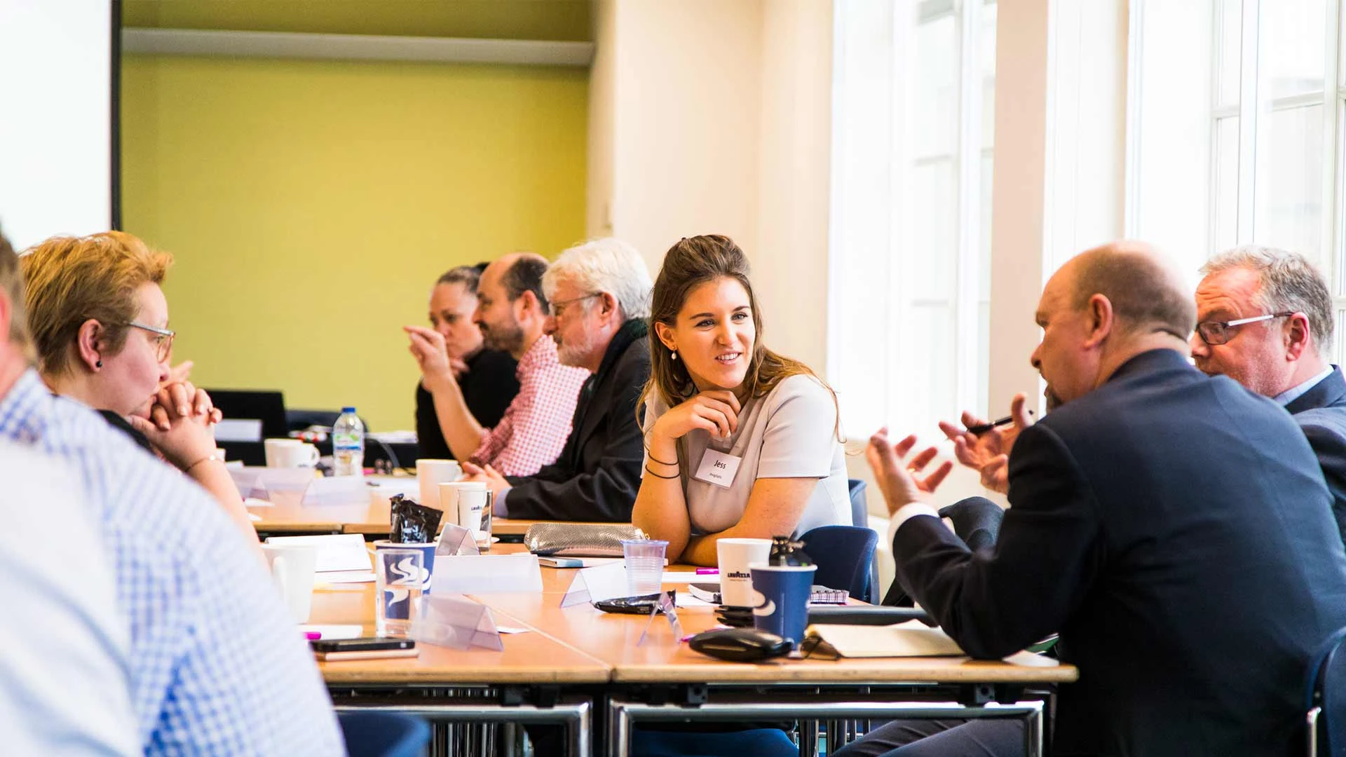 Group of people engaged in discussion around conference tables with papers and coffee cups.