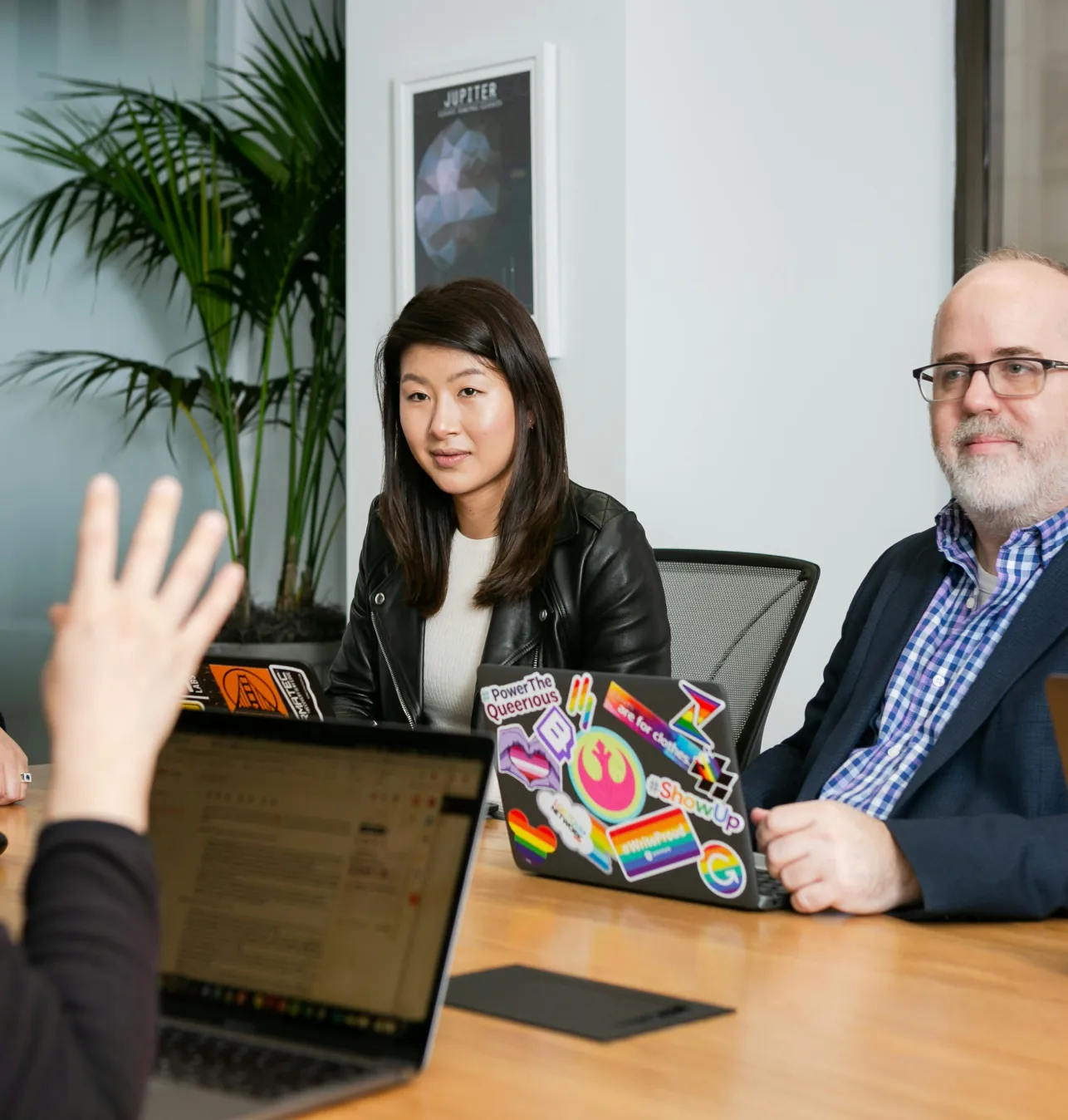 Two people sitting at a conference table with laptops, one listening attentively while another person gestures with a raised hand.