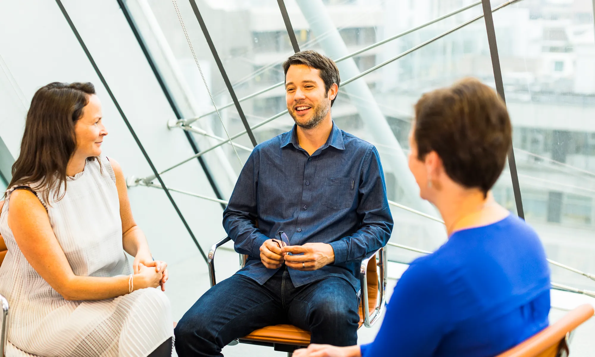 Three people sitting in chairs having a friendly conversation in a bright modern office space with large glass windows.
